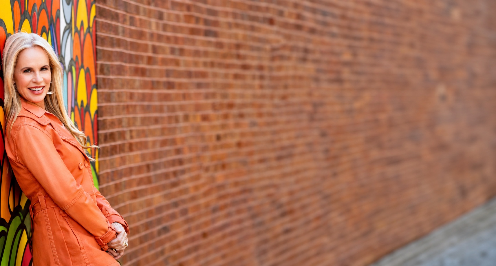 Elisabeth Leamy in orange beside a colorful mural and a long brick wall.